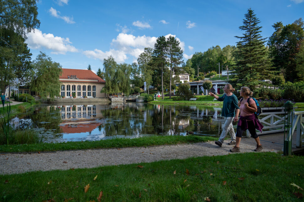 Two people walk along the pond in the spa park, the festival hall can be seen in the background